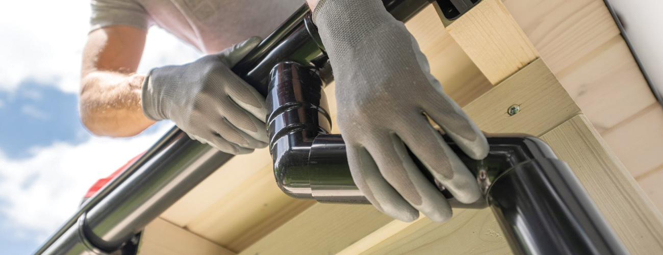 Worker in red cap performing gutter repair on a house, adjusting black metal downspouts while standing on a ladder.